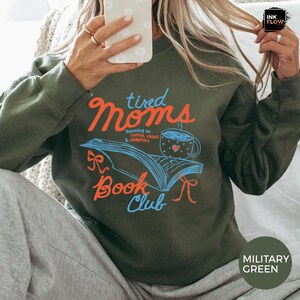 May include: Military green sweatshirt with the text "tired Moms Book Club" in orange and blue. The design includes an open book, a coffee cup, and the phrase "Running on coffee, chaos & chapters."