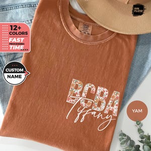Op de afbeelding: Een roestkleurig T-shirt met een bloemenpatroon dat "BCBA Tiffany" in witte tekst spelt. Het shirt is gemaakt van een zacht, comfortabel materiaal en is verkrijgbaar in verschillende kleuren.