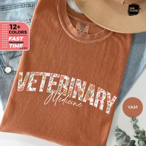 May include: A brown t-shirt with the text "Veterinary Medicine" in white letters surrounded by a floral pattern. The shirt is made of a comfortable, soft fabric.