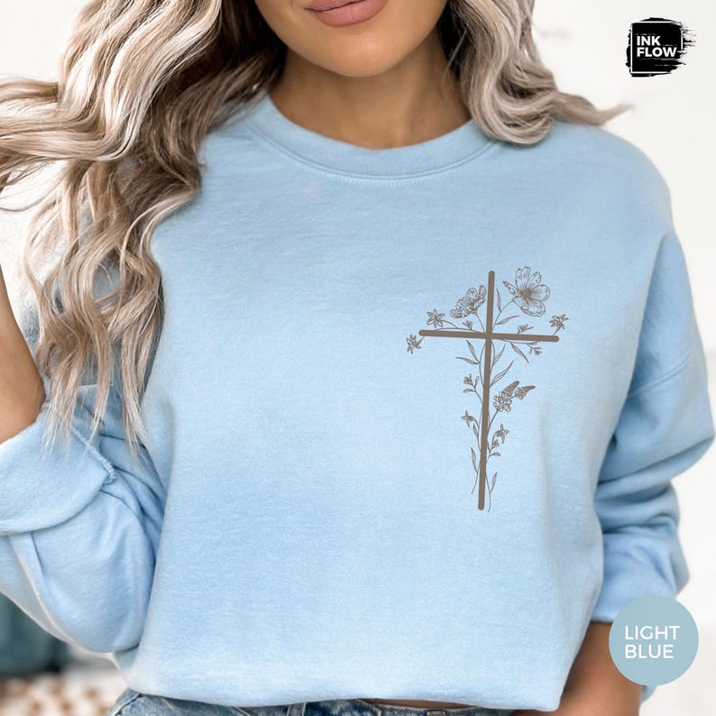 May include: Pale blue sweatshirt featuring a floral cross design. The cross is composed of lines and decorated with flowers and leaves. The text "LIGHT BLUE" is in the bottom right corner.