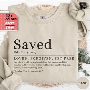 May include: A sand-coloured sweatshirt with the word "Saved" printed in black. The definition below reads "LOVED, FORGIVEN, SET FREE." Additional text explains the meaning. A graphic on the left indicates 12+ colours and fast processing.