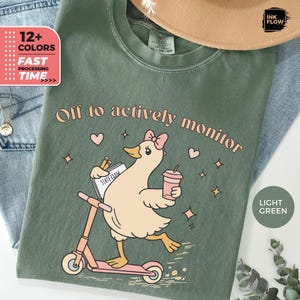 Può includere: T-shirt verde chiaro con un'anatra dei cartoni animati su un monopattino rosa, che tiene in mano una bevanda e un libro con la scritta "STATE EXAM". Il testo recita "Off to actively monitor". Ulteriori testi indicano "12+ COLORI" e "FAST PROCESSING TIME".