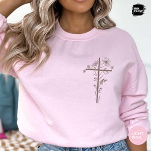 May include: Pale pink sweatshirt with a brown cross design and floral details. The top has a crew neck and long sleeves. The words "LIGHT PINK" are printed on the bottom right corner.