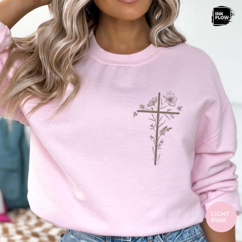 May include: Pale pink sweatshirt with a brown cross design and floral details. The top has a crew neck and long sleeves. The words "LIGHT PINK" are printed on the bottom right corner.