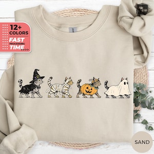 Puede incluir: Una sudadera de cuello redondo color arena con un estampado gráfico de cuatro gatos atigrados con disfraces de Halloween. Los gatos están vestidos de bruja con sombrero negro, momia con vendas blancas, calabaza y fantasma bajo una sábana.