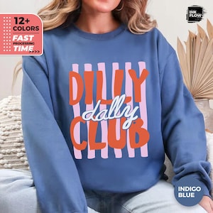 May include: An indigo blue crewneck sweatshirt featuring a graphic design with vertical light pink stripes. The text "DILLY" and "CLUB" is printed in bold orange letters, while the word "dally" is written in white script across the center.