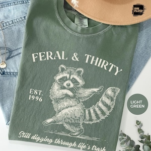 May include: Light green t-shirt with the text "FERAL & THIRTY" and a raccoon graphic. The shirt also features "EST. 1996" and the phrase "Still digging through life's trash." A tan hat and blue jeans are in the background.
