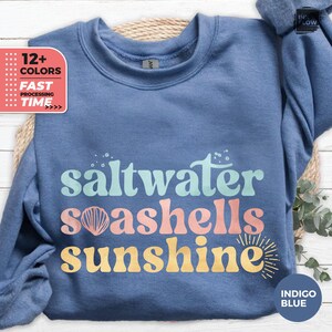 以下が含まれることがあります： インディゴブルーのスウェットシャツ。ターコイズ、コーラル、イエローで「saltwater seashells sunshine」の文字がプリントされています。スウェットシャツはクルーネックと長袖です。画像には「12+色」と「高速処理時間」の文字も含まれています。
