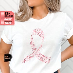 Könnte beinhalten: Weißes T-Shirt mit einem rosafarbenen Banddesign aus kleineren rosafarbenen Bändern und Herzen. Das Band trägt die Worte "Hoffnung", "Stärke", "Heilung", "Kampf" und "Glaube".
