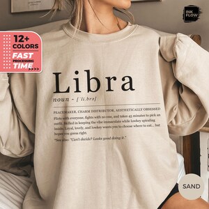 Könnte beinhalten: Sandfarbenes Sweatshirt mit dem Wort "Libra" in großen schwarzen Buchstaben. Darunter befindet sich eine Definition und Beschreibung des Sternzeichens Waage. Das Sweatshirt hat auch eine Grafik mit der Aufschrift "12+ FARBEN" und "SCHNELLE BEARBEITUNGSZEIT".
