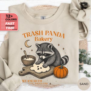 May include: A sand-colored crewneck sweatshirt featuring a graphic of a raccoon kneading dough on a wooden board. The text reads TRASH PANDA Bakery and WE KNEAD EM YOU NEED EM. A bowl, a crescent moon, and an orange pumpkin are also illustrated.