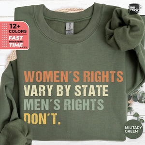 May include: An olive green crewneck sweatshirt featuring the text "WOMEN'S RIGHTS VARY BY STATE MEN'S RIGHTS DON'T." The lettering is displayed in four horizontal lines using orange, cream, and light green colors with a distressed texture.