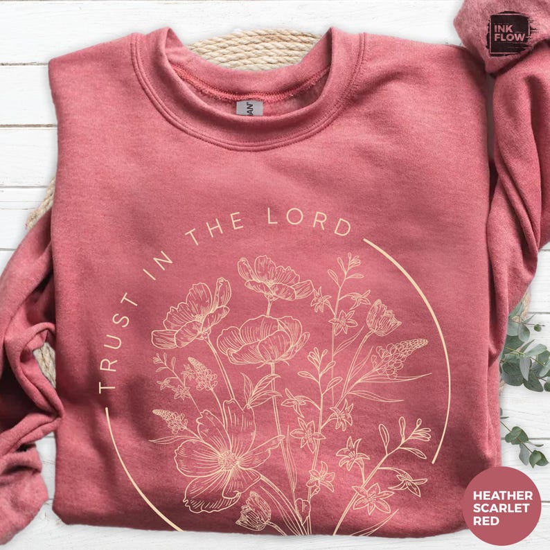 May include: Heather scarlet red sweatshirt with the text "TRUST IN THE LORD" and a floral design in a circular shape. The design features various flowers and foliage in a light beige color. The sweatshirt is made of a soft material.