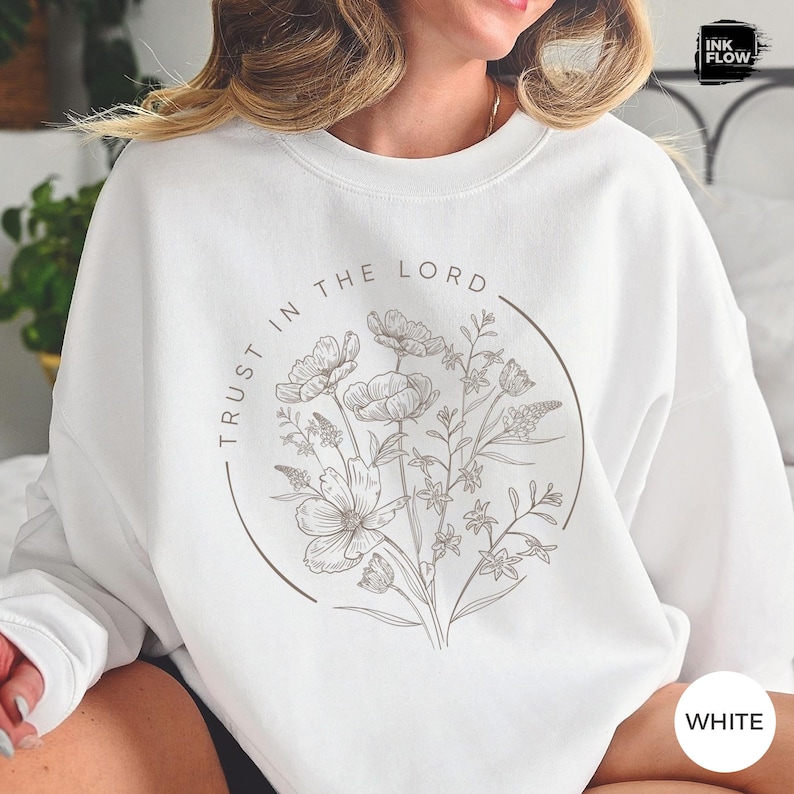 May include: White sweatshirt featuring a floral design with the text "TRUST IN THE LORD" in a circular frame. The floral illustration includes various flower and leaf shapes in a neutral color. The sweatshirt has a crew neck.