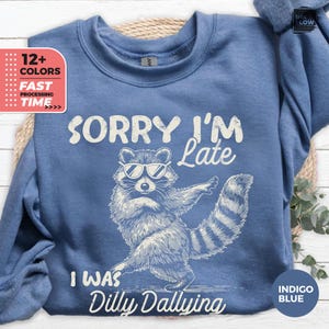 May include: Indigo blue sweatshirt with the text "SORRY I'M Late" and "I WAS Dilly Dallying". The graphic features a raccoon wearing sunglasses. The sweatshirt has a crew neck and long sleeves.