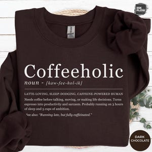 Può includere: Felpa color cioccolato fondente con la parola "Coffeeholic" in grandi lettere bianche. Sotto è stampata la definizione di "Coffeeholic", che descrive un essere umano alimentato dalla caffeina. La felpa è del marchio Gildan.