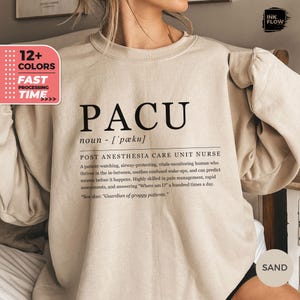 Puede incluir: Una sudadera beige con el texto "PACU" y una definición del acrónimo: "POST ANESTHESIA CARE UNIT NURSE". La definición describe en detalle el papel de una enfermera de PACU.