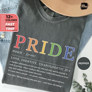 May include: A dark gray t-shirt with the word "PRIDE" in rainbow colors. The shirt includes a definition and a message about love, identity, and joy. The shirt is part of a collection with 12+ colors and fast processing.