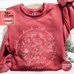 Peut inclure: Sweat-shirt rouge écarlate chiné avec un motif floral et le texte "He leads me beside still waters, He restores my soul." Le motif comprend une couronne florale avec le texte en cercle. Le sweat-shirt a également le texte "12+ COLORS" et "FAST PROCESSING TIME."