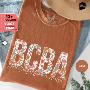Op de afbeelding: Een roestkleurig T-shirt met een bloemenontwerp dat "BCBA" spelt en de tekst "Board Certified Behavior Analyst" eronder.