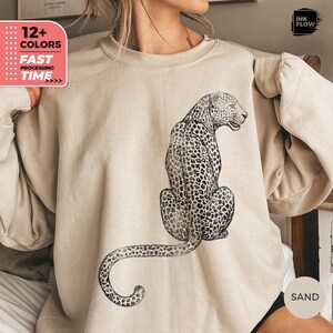 May include: A sand-colored crewneck sweatshirt featuring a black ink-style illustration of a leopard sitting in profile. The leopard is depicted with detailed spots and a long, curved tail extending toward the bottom hem of the garment.
