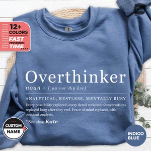 Pode incluir: Camisola azul índigo com a palavra "Overthinker" em branco e sua definição. A camisola tem gola redonda e mangas compridas. A imagem também inclui texto que diz "12+ CORES" e "TEMPO DE PROCESSAMENTO RÁPIDO".