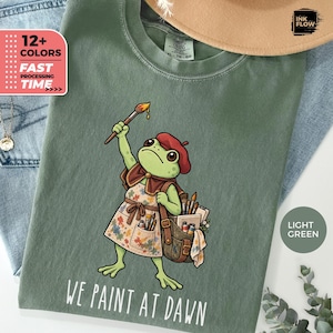 May include: A light green t-shirt featuring a graphic of a frog wearing a red beret and a paint-splattered apron. The frog holds a paintbrush and carries a satchel filled with art supplies. The text below the frog reads, WE PAINT AT DAWN.