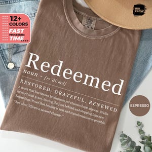 Könnte beinhalten: Braunes T-Shirt mit dem weißen Aufdruck "Redeemed" sowie dessen Definition und zugehörigem Text. Das Bild enthält eine rosa Box mit "12+ Farben" und "Schnelle Bearbeitungszeit". Die T-Shirt-Farbe ist Espresso.