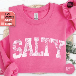May include: A bright pink sweatshirt with the word "SALTY" in white, featuring a palm tree leaf design within the letters. The image also includes text that says "12+ COLOURS" and "FAST PROCESSING TIME".