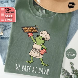 May include: A light green t-shirt featuring a graphic of a green frog wearing a white chef hat and apron, holding a baking sheet with chocolate chip cookies. The text "WE BAKE AT DAWN" is printed in white below the frog illustration.