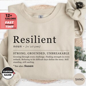 May include: A sand-colored crewneck sweatshirt featuring black text that defines the word Resilient as a noun, followed by the descriptors strong, grounded, unbreakable, and a paragraph about growing through challenges, ending with see also Susan.