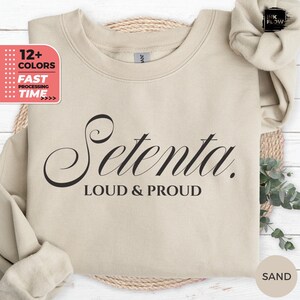 May include: A sand-colored sweatshirt with the word "Setenta" in a stylish black font, with "LOUD & PROUD" below. The image also shows a scrunchie and a graphic with "12+ COLORS" and "FAST PROCESSING TIME".