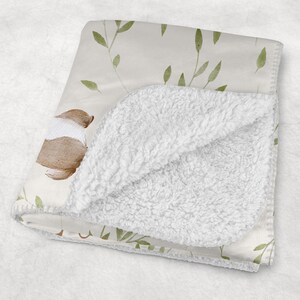 May include: A folded baby blanket with a cream-colored floral pattern and a soft, white sherpa lining. The blanket features a watercolor design of brown and white flowers and green leaves.