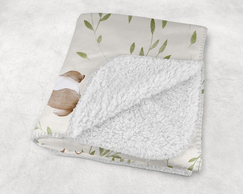 May include: A folded baby blanket with a cream-colored floral pattern and a soft, white sherpa lining. The blanket features a watercolor design of brown and white flowers and green leaves.