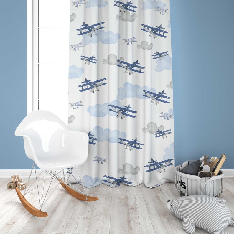 May include: White window curtains with a repeating pattern of blue airplanes and clouds. The curtains are hanging in front of a window, with a white rocking chair and wooden floor in the foreground. The curtains add a playful touch to the room.