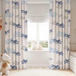 May include: Cream-colored nursery curtains with a repeating pattern of blue biplanes and light blue clouds. The curtains are hanging in front of a window, with a white window shade. The room has a neutral color scheme.
