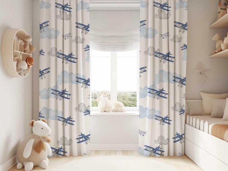 May include: Cream-colored nursery curtains with a repeating pattern of blue biplanes and light blue clouds. The curtains are hanging in front of a window, with a white window shade. The room has a neutral color scheme.