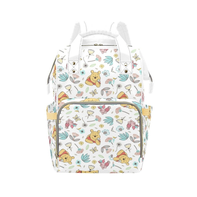 May include: White diaper bag with a Winnie the Pooh and Piglet print. The bag has a floral pattern with butterflies and leaves. The bag has a top handle and adjustable shoulder straps.