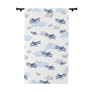 May include: White fabric shower curtain featuring a repeating pattern of blue biplanes and light blue clouds. The design includes gray clouds and smaller, single-wing planes. The curtain is hung on a black rod.