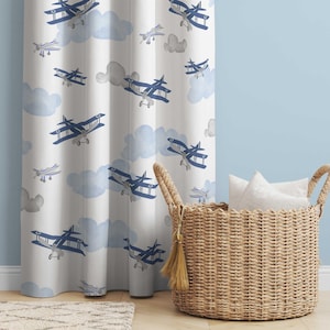 May include: White curtain with a pattern of blue biplanes and light blue clouds. A woven basket with pillows sits to the right of the curtain. The curtain is hanging in front of a light blue wall.