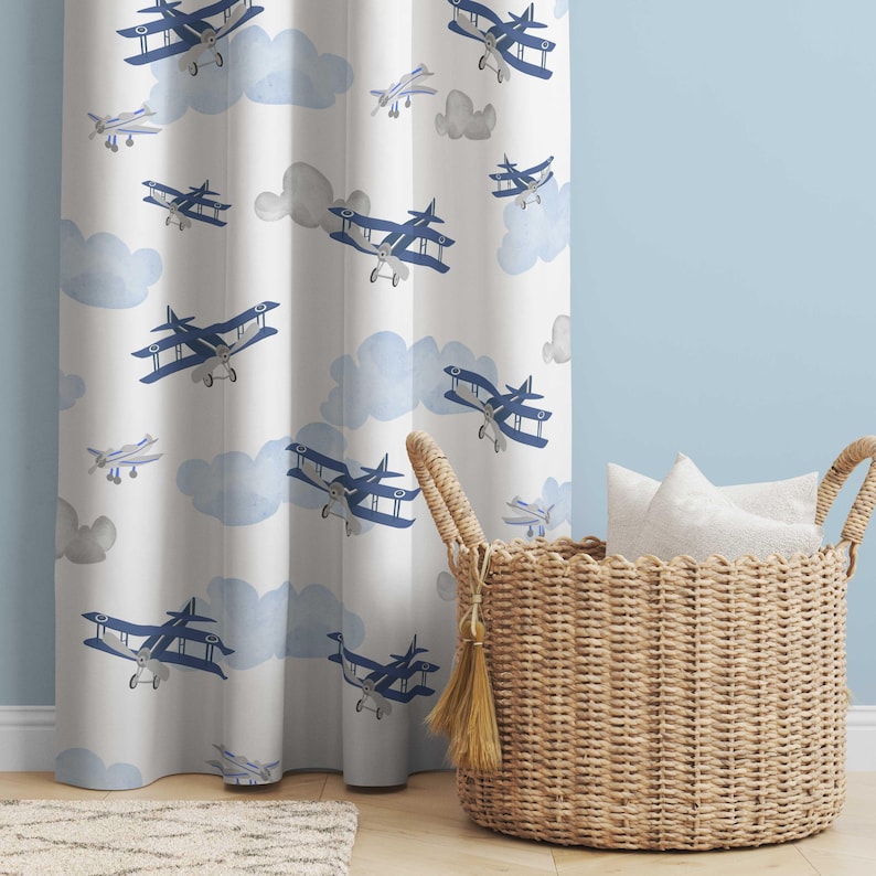 May include: White curtain with a pattern of blue biplanes and light blue clouds. A woven basket with pillows sits to the right of the curtain. The curtain is hanging in front of a light blue wall.