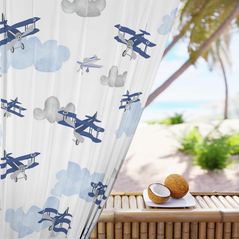 May include: White sheer curtain with a repeating pattern of blue airplanes and light blue clouds. The curtain is partially covering a window with a view of a beach and palm trees. A coconut and a half coconut are on a white plate.