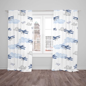 May include: White window curtains with a repeating pattern of blue airplanes and light blue clouds. The curtains are hanging in front of a window with a view of a city skyline.