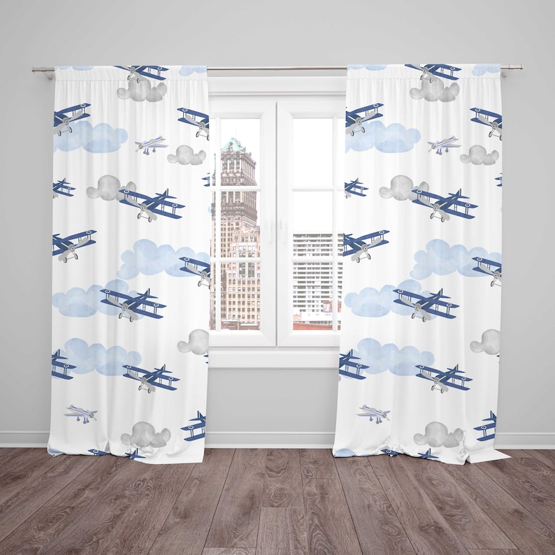 May include: White window curtains with a repeating pattern of blue airplanes and light blue clouds. The curtains are hanging in front of a window with a view of a city skyline.