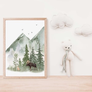 May include: A framed watercolor print featuring a forest landscape with green mountains, tall pine trees, a moose, and a small deer. Above the mountains, four small black birds are flying. Beside the frame is a white crocheted bear toy.