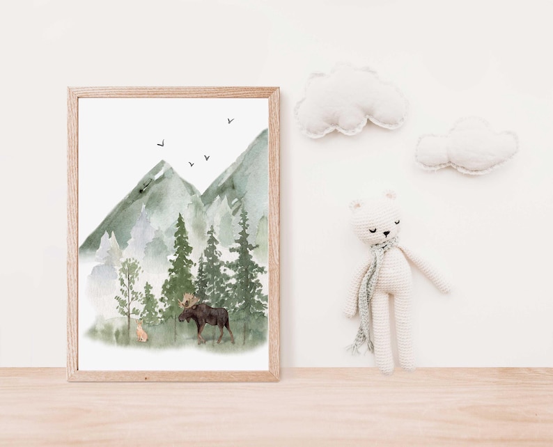 May include: A framed watercolor print featuring a forest landscape with green mountains, tall pine trees, a moose, and a small deer. Above the mountains, four small black birds are flying. Beside the frame is a white crocheted bear toy.