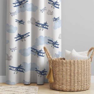 May include: White curtain panel featuring a repeating pattern of blue biplanes and light blue watercolor clouds. A woven basket with white pillows sits to the right of the curtain. The curtain is hanging in front of a light gray wall.