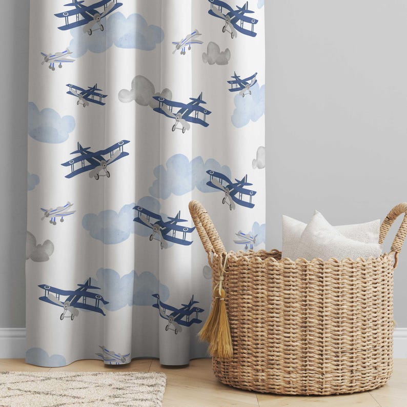 May include: White curtain panel featuring a repeating pattern of blue biplanes and light blue watercolor clouds. A woven basket with white pillows sits to the right of the curtain. The curtain is hanging in front of a light gray wall.