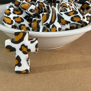 May include: A bowl filled with small, white and brown cross-shaped charms with a leopard print design. One cross charm is sitting in front of the bowl.