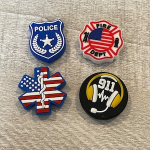 May include: Four colorful, rubber charms with American flag designs. The charms feature a police badge, a fire department logo, a medical symbol, and a 911 emergency symbol.
